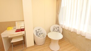 Una habitación pequeña y ordenada con un escritorio, dos sillas, una mesita y una ventana con cortinas traslúcidas.