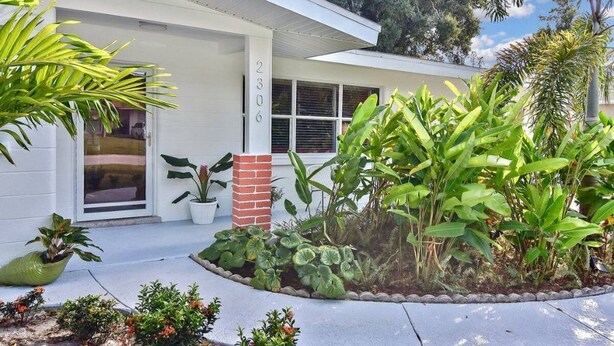 Una casa moderna con fachada blanca, un pilar de ladrillo y un jardín bien cuidado con diversas plantas y un pequeño estanque.