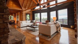 A spacious living room with a white sofa, a coffee table, and a fireplace. Large windows offer a view of the outdoors. - The Ridge: A Modern Cabin (Bostic)