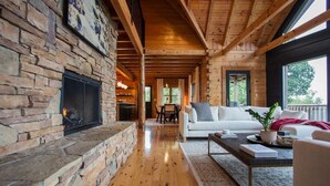 A cozy living room with a stone fireplace, wooden beams, a white sofa, and a large window. - The Ridge: A Modern Cabin (Bostic)