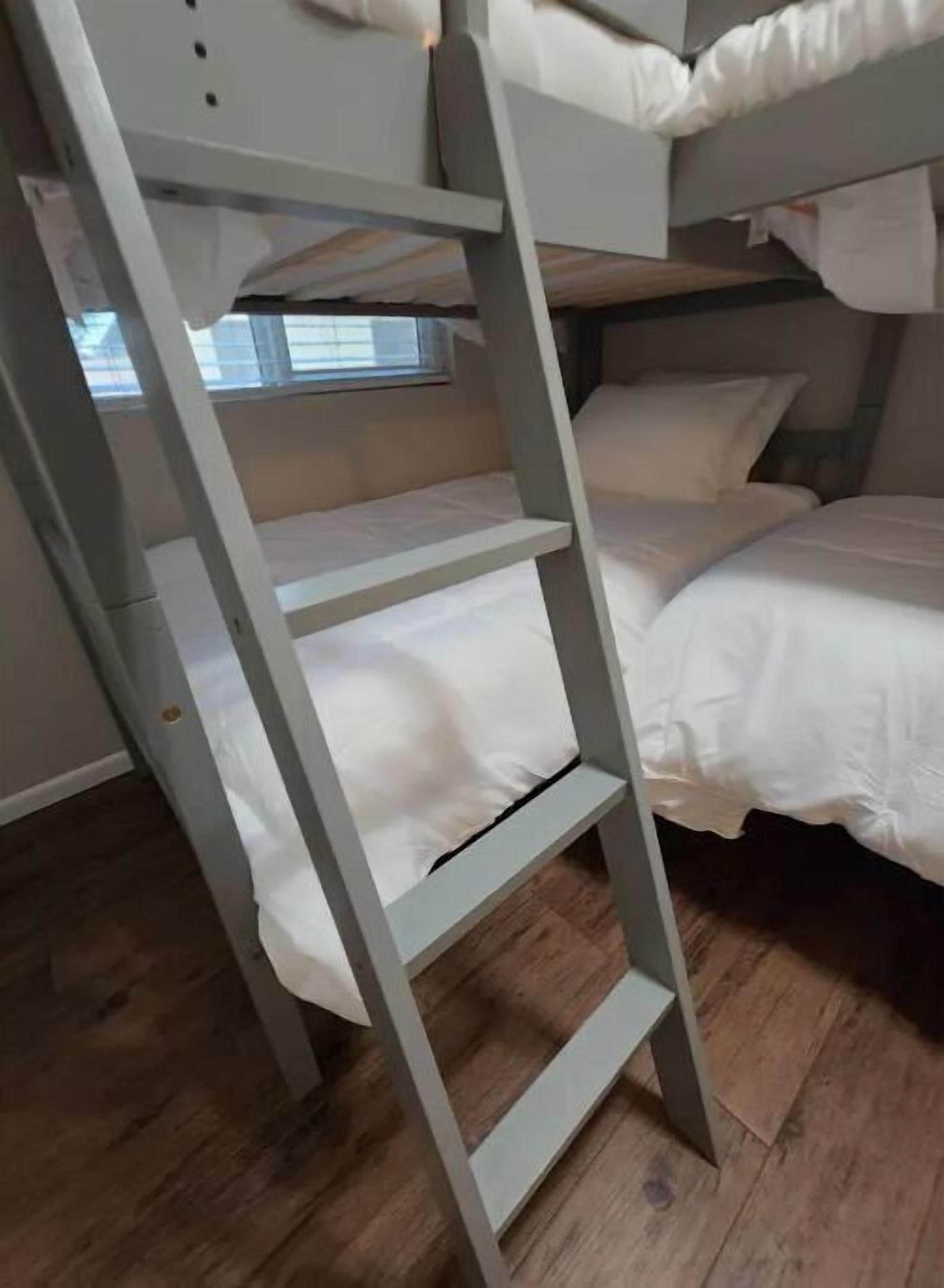 Un lit superposé avec une échelle, du linge de lit blanc et une fenêtre avec des stores.