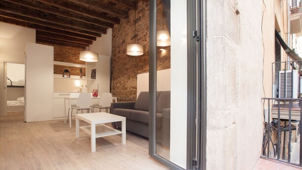A modern interior with a wooden ceiling, a dining area with a white table and chairs, a grey sofa, and a kitchen area with a white countertop and shelves. - Alissia 1 - Habitat Apartments (Barcelona)