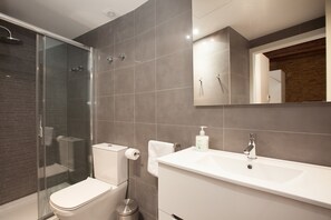A modern bathroom with a glass shower enclosure, a white toilet, a sink with a mirror above it, and a toilet paper roll on the wall.