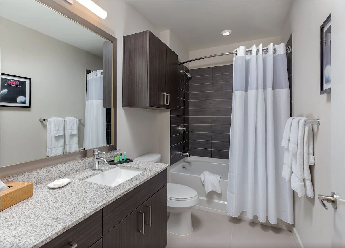 A modern bathroom with a granite countertop sink, a mirror, a dark wood cabinet, a white toilet, a bathtub with a shower curtain, and a towel rack.