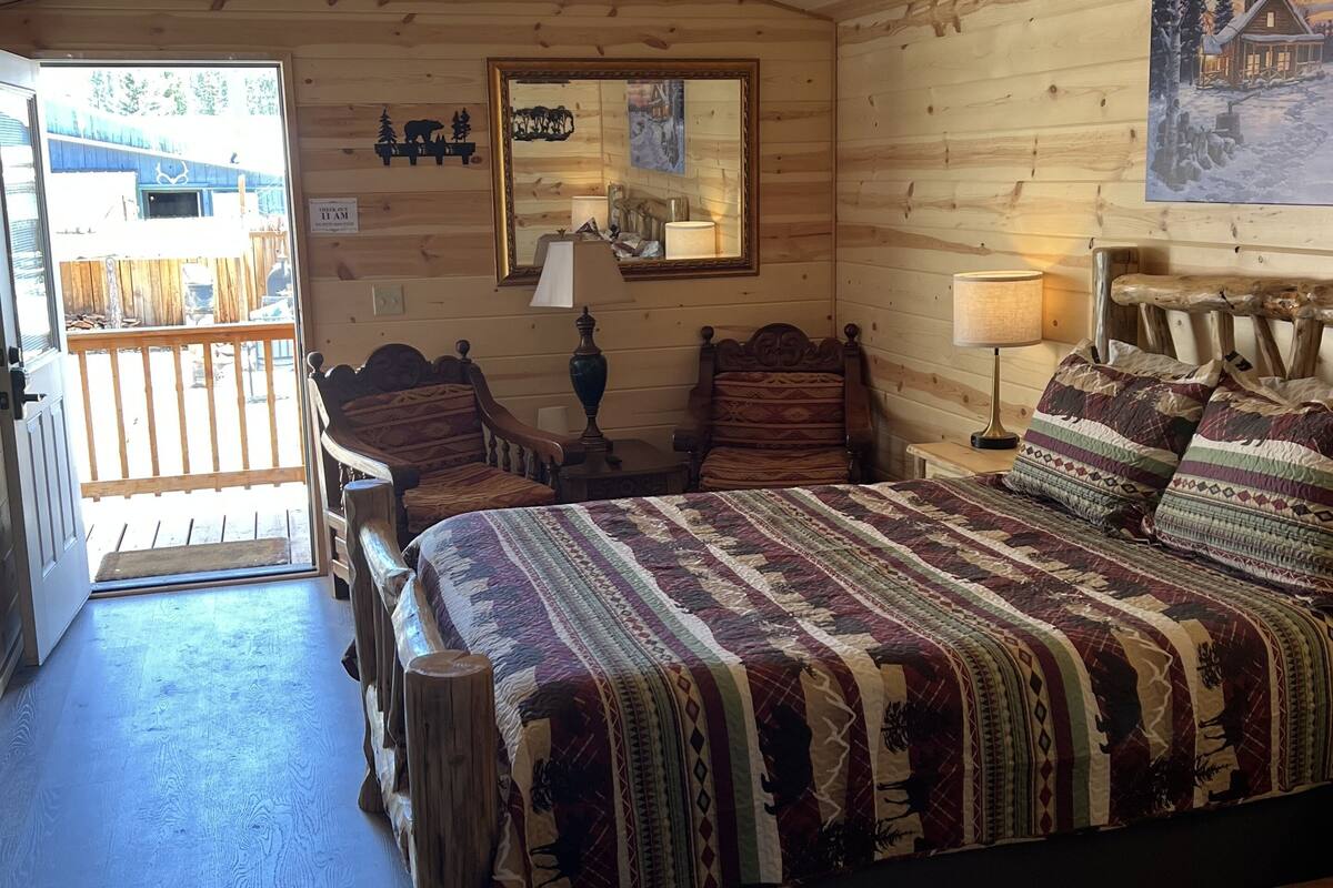 A wooden cabin bedroom with a large bed, two chairs, a wooden headboard, a mirror, and a painting on the wall.