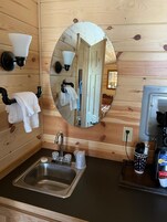 Baño de cabaña de madera con lavamanos, espejo, toallero y un ventanal pequeño.