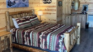 A cozy log cabin bedroom with a wooden bed, a nightstand, a dresser, and a mirror. - The Gold Mine Hotel (Idaho City)