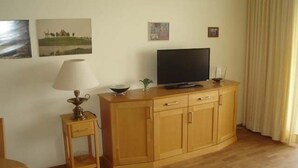 A room with a wooden cabinet, a television on top, a small table with a lamp, and two framed pictures on the wall. - Nautilus, Ferienwohnung 23 (Cuxhaven)