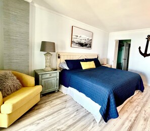 A bedroom with a bed, a nightstand, a lamp, a chair, and a door. - Experience Spectacular Oceanfront Views in our Cozy Condo  (North Myrtle Beach)