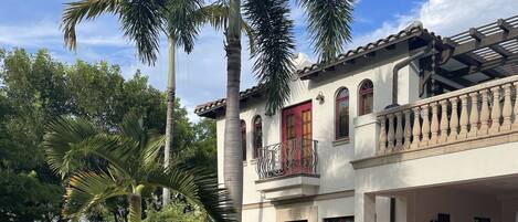 A two-story building with a red door and balcony, surrounded by tropical plants and a paved driveway.
