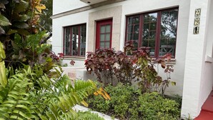 Ein weißes Gebäude mit roten Fensterrahmen, ein kleiner Garten mit verschiedenen Pflanzen und eine rote und weiße Nummernschild.