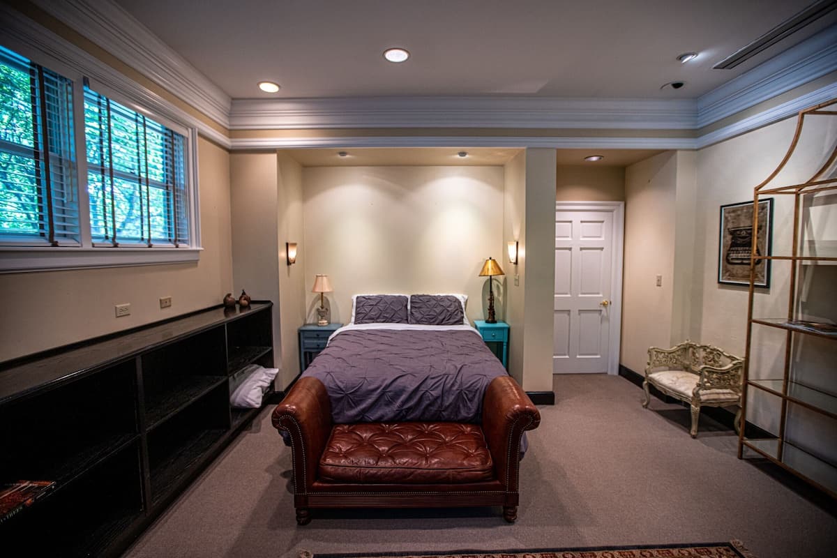 A bedroom with a bed, a bench, a nightstand, a lamp, a chair, and a shelf.