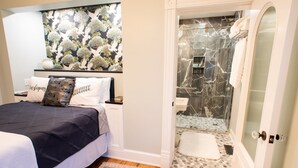 A bedroom with a bed, pillows, a nightstand, a decorative wall, and a bathroom with a shower and glass door. - Washington Street Quarters (Lewisburg)