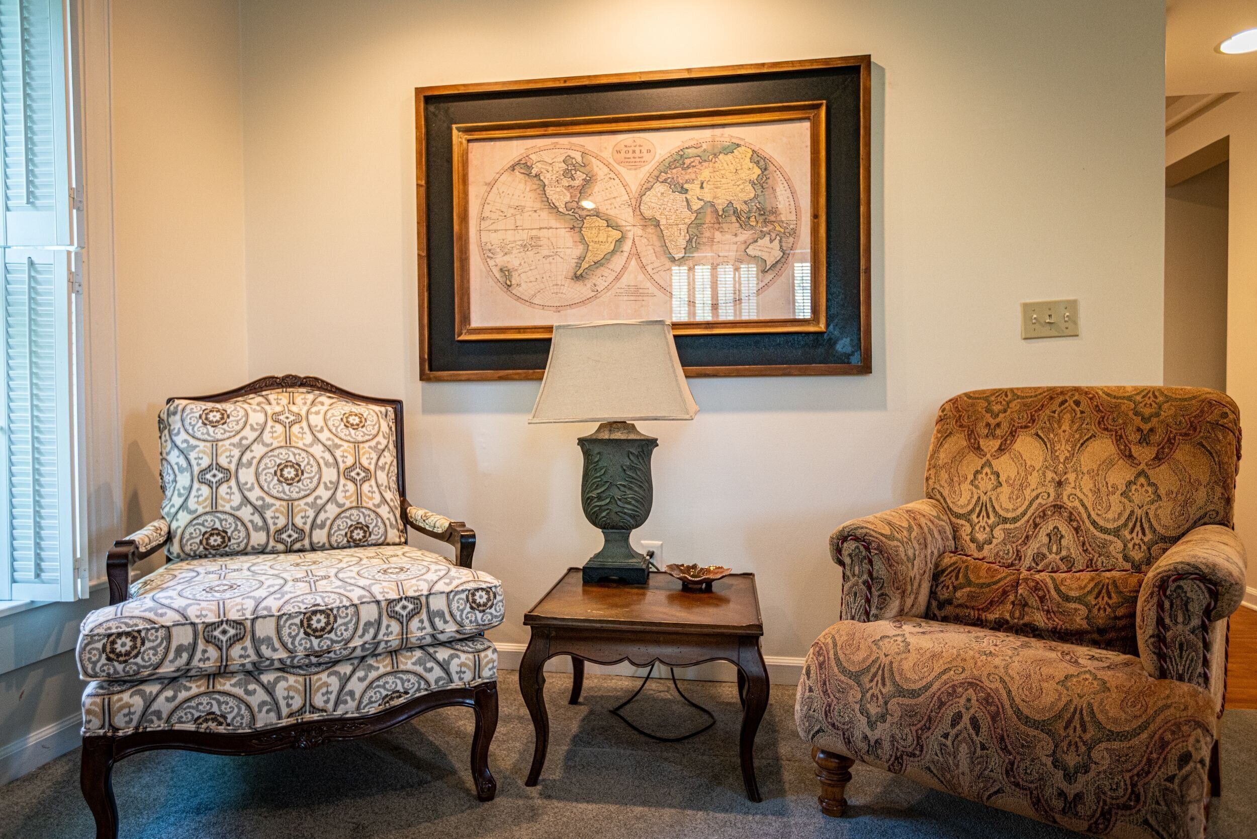Una habitación con dos sillones estampados, una mesita con lámpara y un mapa del mundo enmarcado en la pared.