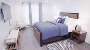 Moderni hotellihuone, jossa on suuri sänky, yöpöytä lampun kanssa, tuoli, työpöytä ja seinälle asennettu televisio.