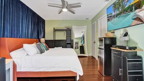 Habitación de hotel con cama, ventilador de techo, minibar y vistas a la zona de la piscina.