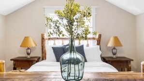 Un dormitorio con cama, dos mesitas de noche con lámparas, una mesa de madera con un florero y un ventanal con vista a una estructura de madera en el exterior.
