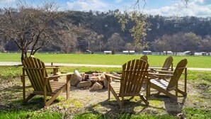 Un grupo de sillas Adirondack de madera alrededor de un pequeño hogar para fuego en un área verde, con vista a una colina boscosa y edificios a lo lejos.