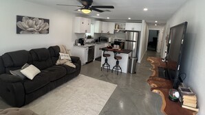 A modern living room with a dark sofa, a white rug, and a kitchen in the background.