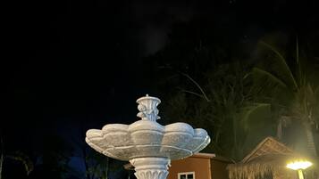 Une scène nocturne mettant en scène une grande fontaine richement décorée dans un jardin, entourée de plantes tropicales et d’un petit bâtiment doté d’un toit de chaume.