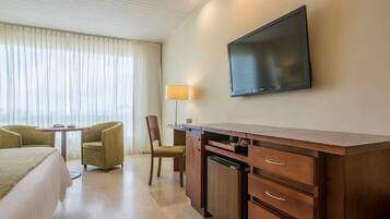 A hotel room with a bed, a desk with drawers, a television mounted on the wall, a lamp, and a chair.