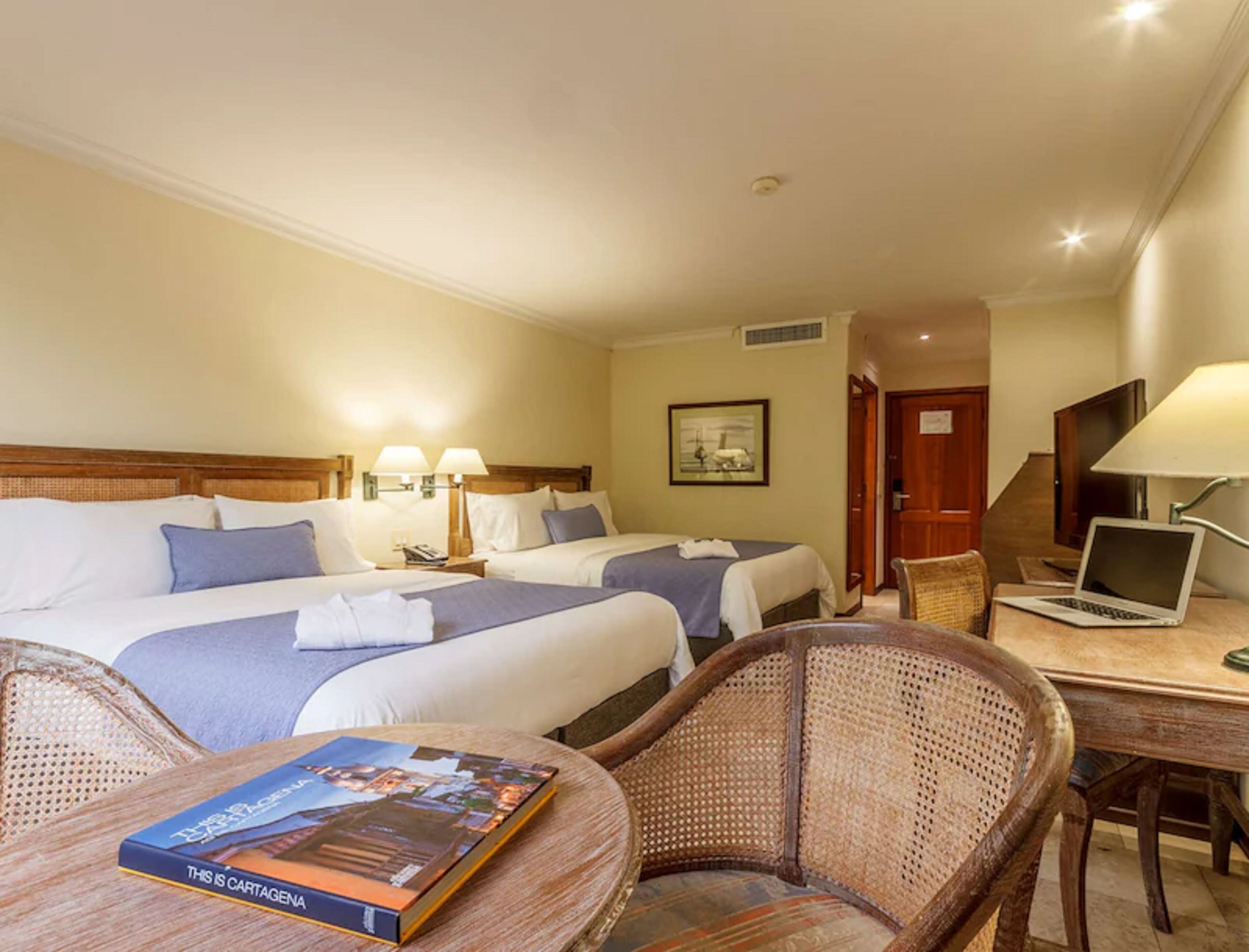 A hotel room with two beds, a desk with a laptop, a wicker chair, and a book on the table.