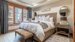 A bedroom with a large bed, a bench, a nightstand, a mirror, and a view of buildings outside. - Premier 1 BD/1 BR condo ski in and out.  Gondola is right outside the condo. (Truckee)