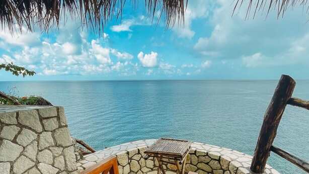 A wooden bench with a red cushion, a small table, and a stone wall overlooking the sea. - Negril Bungalow Paradise overlooking cliffs with Private Snorkeling Entrance. (Hanover Parish)