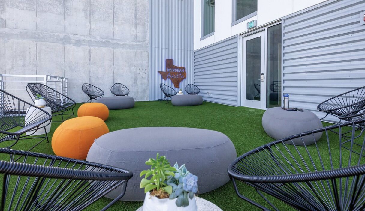 Un espace extérieur aménagé avec des chaises à armature métallique noire et des poufs gris et orange posés sur un gazon synthétique, avec un mur en métal et une enseigne en arrière-plan.