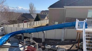 Un jardin avec un toboggan bleu, une terrasse en bois et une maison entourée d’une clôture blanche.