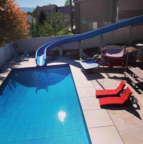 A swimming pool with a blue slide, lounge chairs, and a view of the surrounding area. - Walkout Basement Apt w/ Waterslide, Hot Tub & More
 (South Jordan)