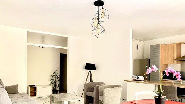 Een moderne woonkamer met een grijze bank, een kleine witte tafel en een rode tafel met stoelen. Er is een keuken met houten kasten en een magnetron.
