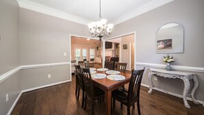 Comedor con una mesa de madera dispuesta para seis personas, rodeada de sillas. Un candelabro cuelga del techo, y un aparador con un jarrón de flores está apoyado en la pared.