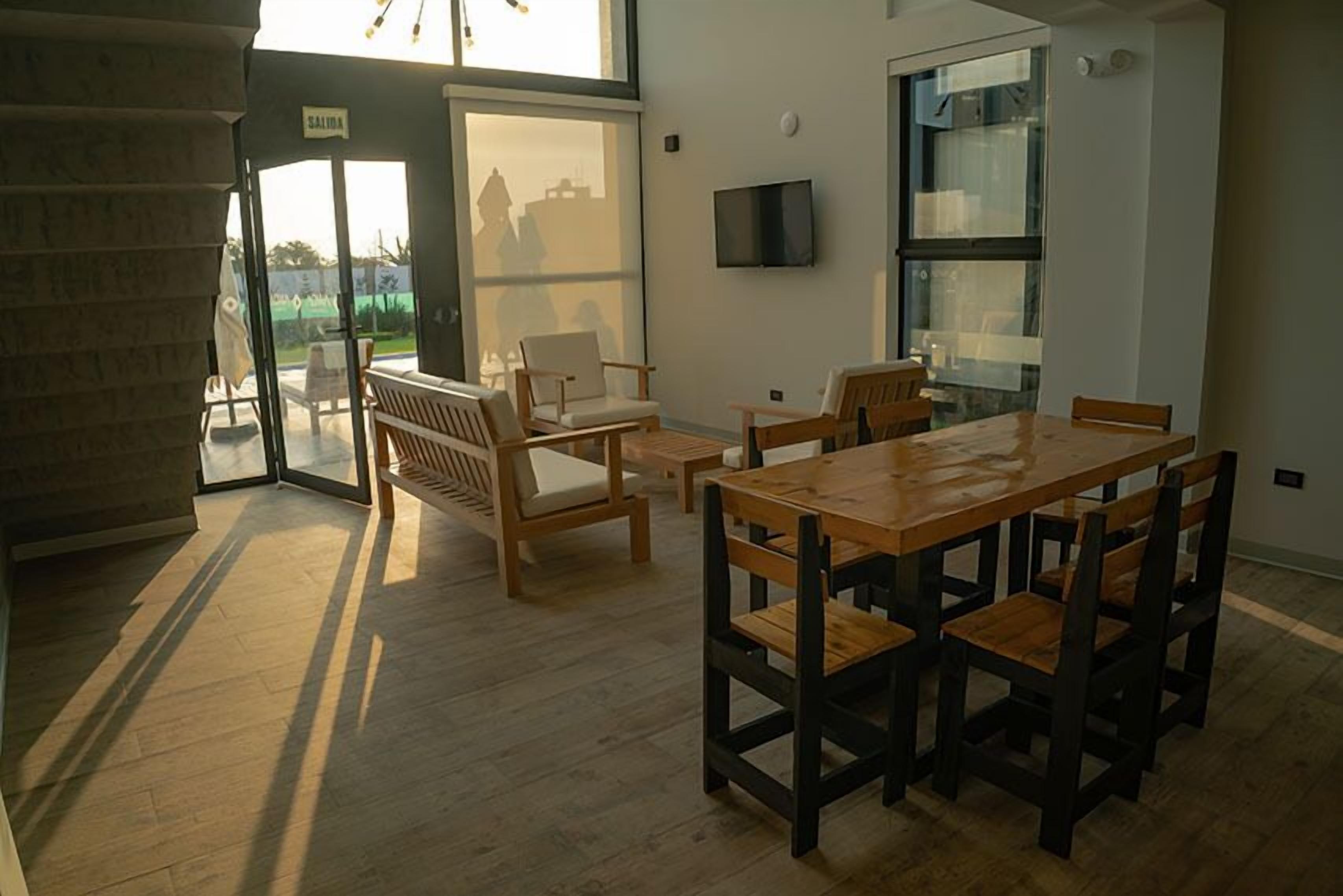 A dining area with a wooden table and chairs, a TV mounted on the wall, and a large window with a view of the outside.