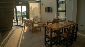 A dining area with a wooden table and chairs, a TV mounted on the wall, and a large window with a view of the outside.