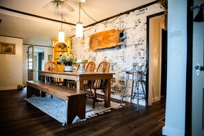 A dining area with a wooden table and chairs, a hanging light fixture, and a brick wall with a wooden board.