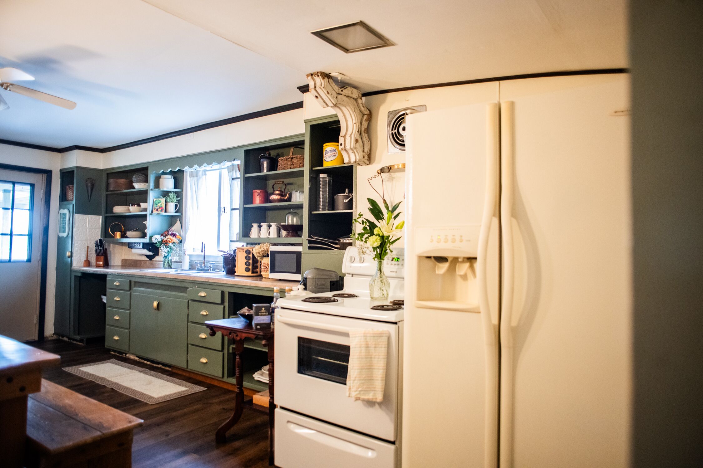 Una cocina con un refrigerador blanco, una estufa y un microondas. En primer plano, hay una mesa de madera y una silla.