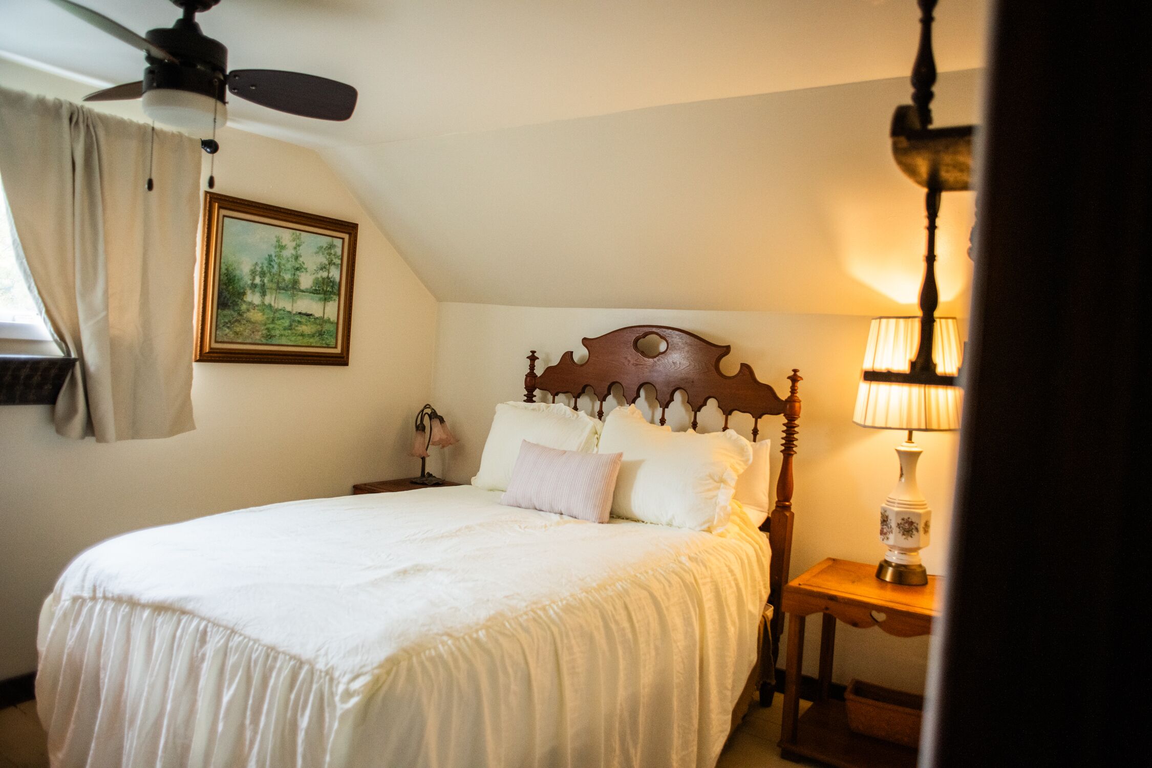 Un dormitorio con cabecera de madera, cama con ropa de cama blanca, mesita de noche con lámpara y un cuadro en la pared.