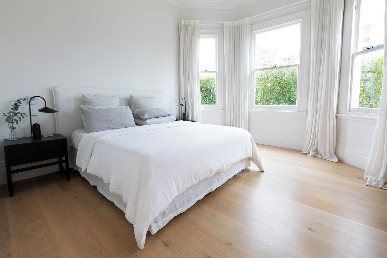 A bedroom with a large bed, white bedding, a bedside table with a lamp, and a window with curtains.