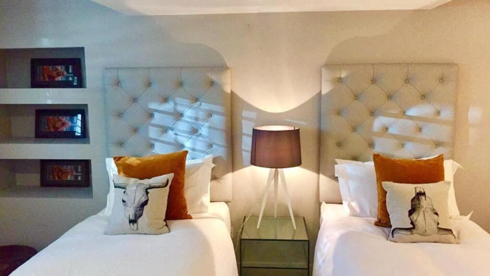 Habitación de hotel con dos camas, cada una con tablero de cabecera capitonado, mesita de noche, lámpara y cojines decorativos.