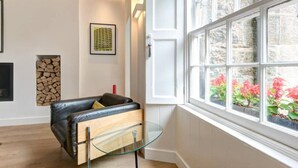 Un salon moderne avec un canapé en cuir, une table basse en verre et une grande fenêtre donnant sur un jardin fleuri et un mur en pierre.