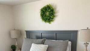 A neatly made bed with a grey bedspread, two grey pillows, and a white pillow. There is a small green wreath on the wall above the bed.