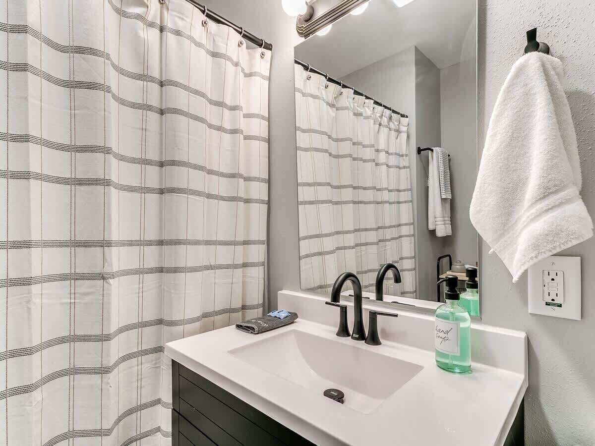 Ein Badezimmer mit einem weißen Waschbecken, einem schwarzen Wasserhahn und einem Spiegel darüber. Ein gestreifter Duschvorhang und ein weißes Handtuch hängen an der Wand.