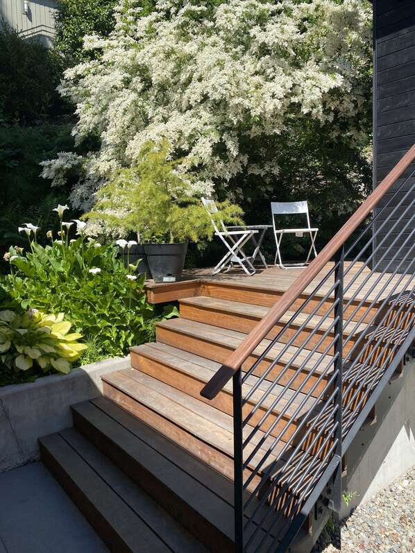 Una terraza de madera con barandilla metálica, rodeada de vegetación exuberante y plantas con flores blancas.