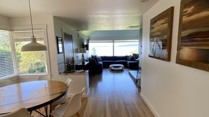 A spacious living area with a wooden floor, a round dining table with chairs, a sofa, and a TV mounted on the wall. - Peaceful View Home with a Cozy Modern feel walking distance to the beach (Seattle)