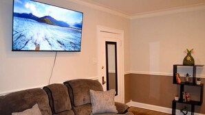 Ett vardagsrum med en stor, platt-TV som är monterad på väggen, en brun soffa med kuddar, ett svart sidobord med dekorativa föremål och ett trägolv.
