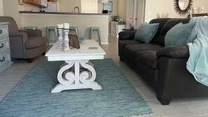 Ein Wohnzimmer mit einem weißen Couchtisch, einem braunen Sofa und einem blauen Teppich. Im Hintergrund ist eine Küche mit Holzschränken und einem Fenster zu sehen.