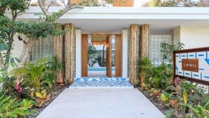 A hotel entrance with a tiled pathway leading to a wooden door, surrounded by tropical plants and a signboard.