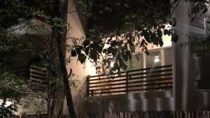 A nighttime view of a building with a wooden balcony, surrounded by dense foliage and a bamboo fence.
