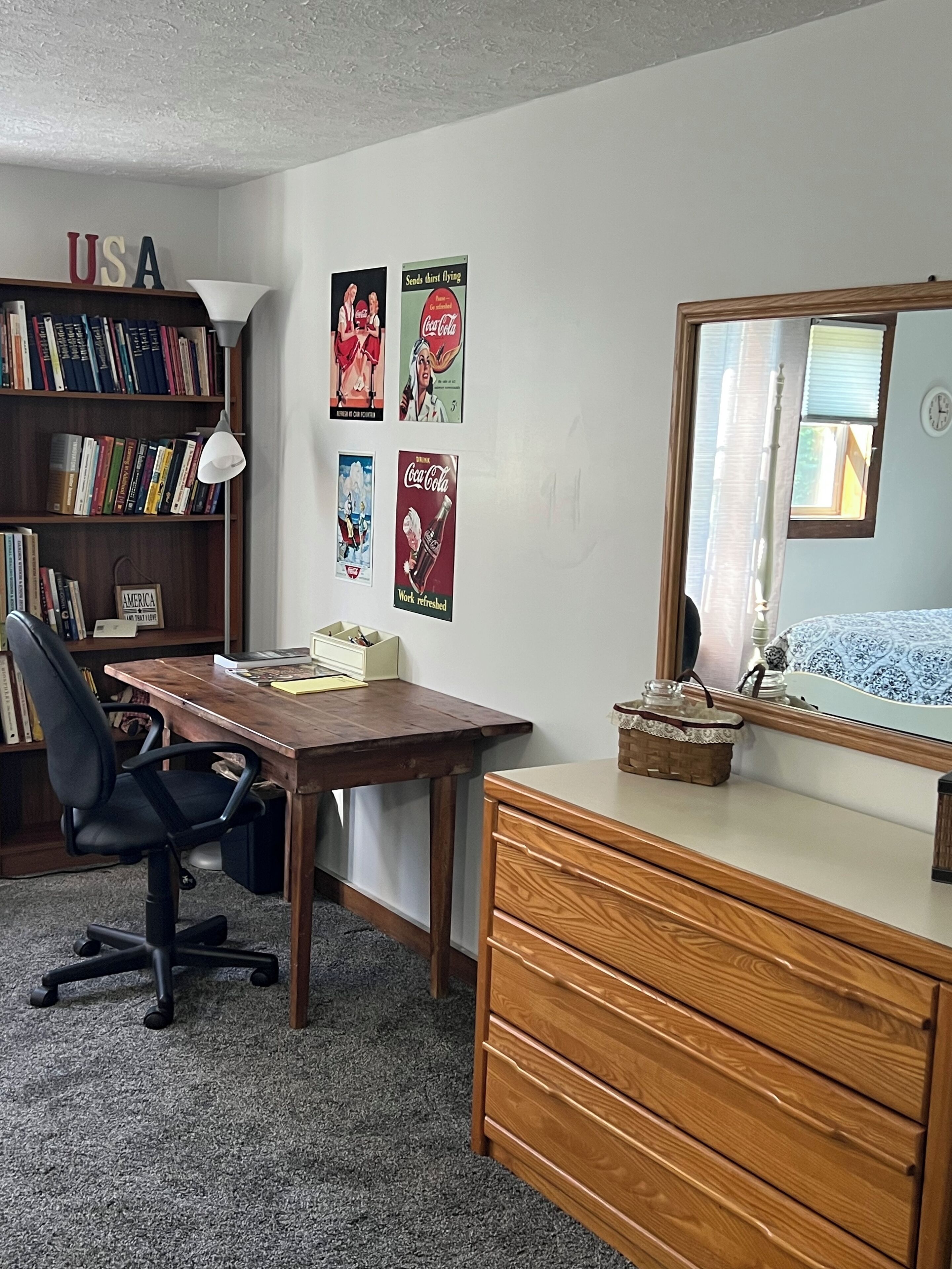 A room with a wooden desk, a chair, a dresser, a bookshelf, and wall decorations including posters.